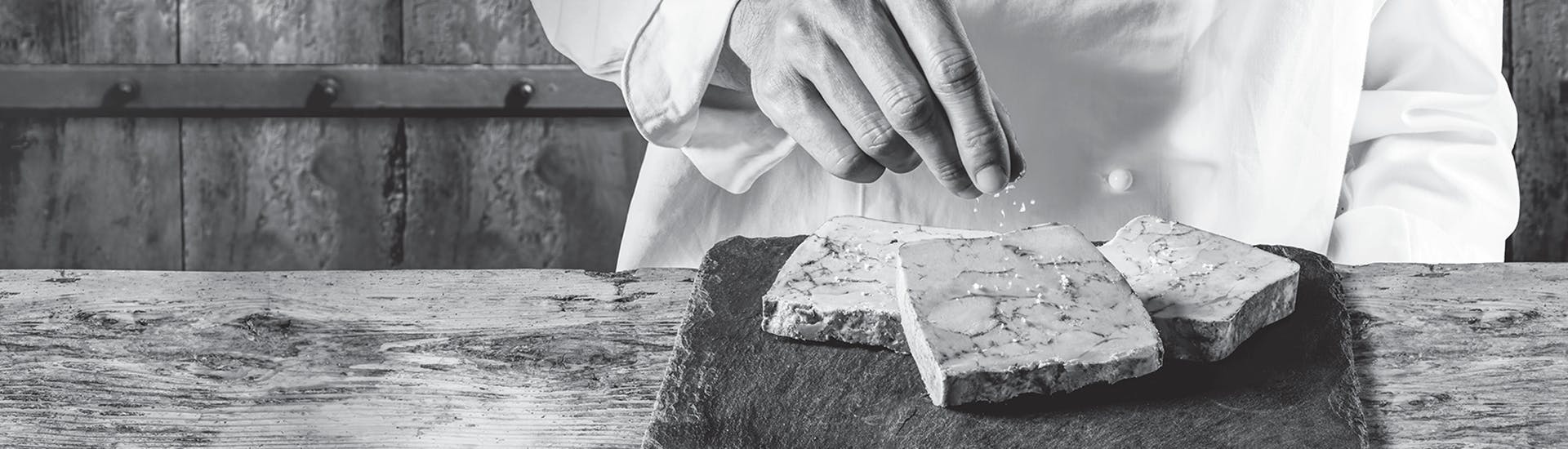 Photo en noir et blanc d'un cuisinier salant le fois gras disposé sur une ardoise posée sur une table en bois.